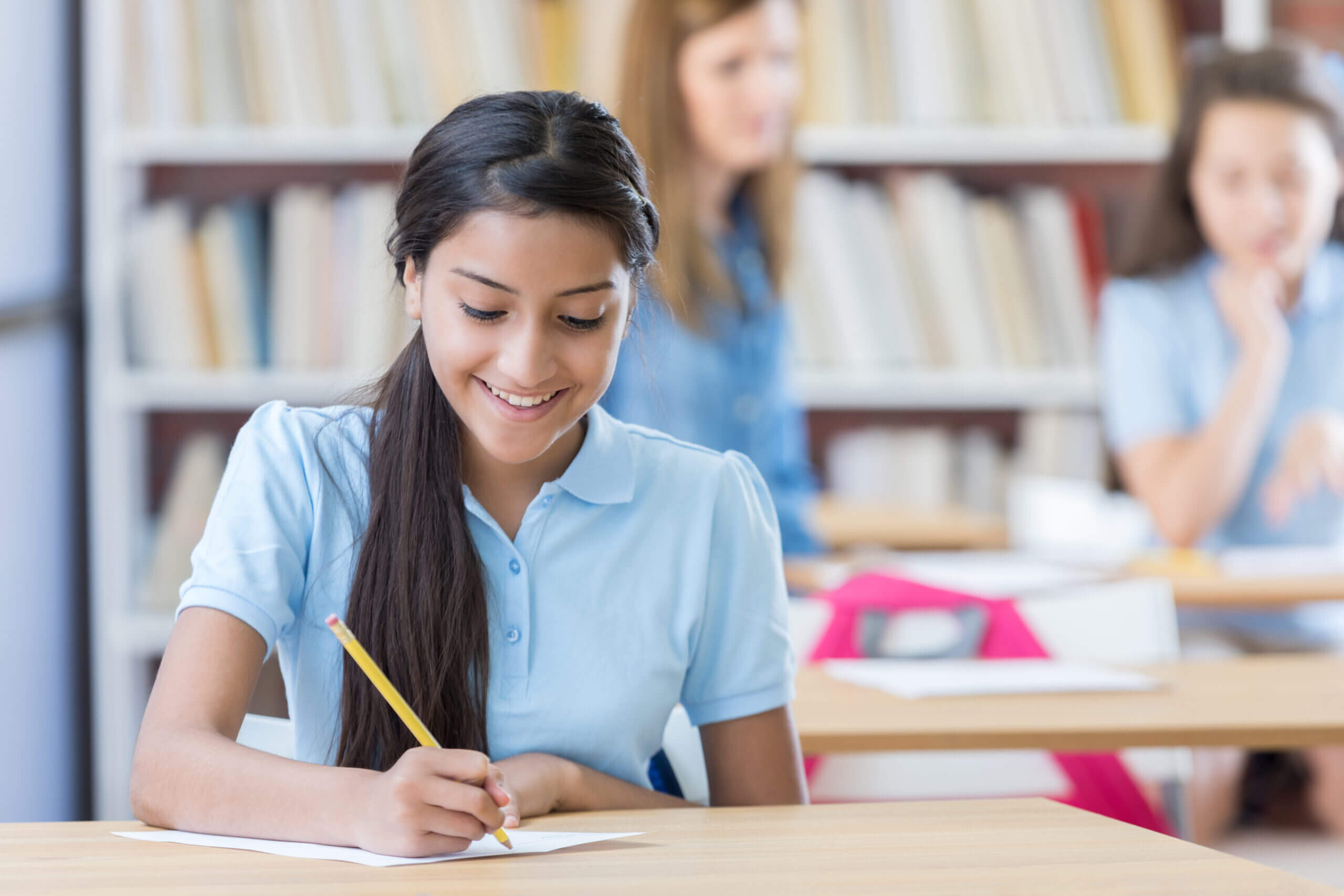Happy Hispanic STEM schoolgirl smiles confidently while working on an assignment in the library or classroom. A teacher is working with a student in the background.
