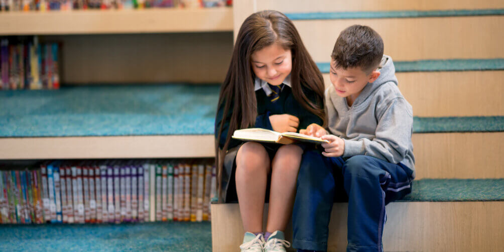 Two students sit as the bottom of the stairs in the library to read to one another.